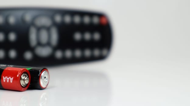 Black TV remote control with AAA alkaline batteries in red and white on a white background. Battery replacement, spare parts. Remote control battery compartment close-up. Copy space.