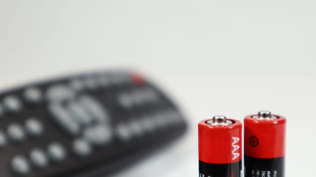 Black TV remote control with AAA alkaline batteries in red and white on a white background. Battery replacement, spare parts. Remote control battery compartment close-up