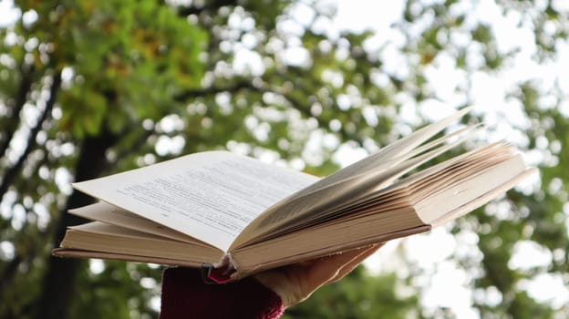 Female hand holding open book with sunlight as concept of freedom of knowledge on green bokeh background in park outdoors. Open book on nature abstract light background