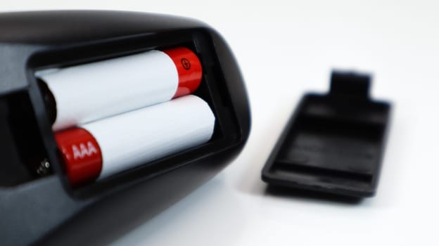Black TV remote control with AAA alkaline batteries in red and white on a white background. Battery replacement, spare parts. Remote control battery compartment close-up