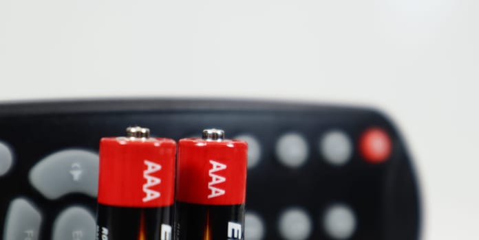 Black TV remote control with AAA alkaline batteries in red and white on a white background. Battery replacement, spare parts. Remote control battery compartment close-up