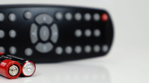 Black TV remote control with AAA alkaline batteries in red and white on a white background. Battery replacement, spare parts. Remote control battery compartment close-up