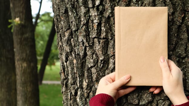 A closed book in a cover made of craft paper in female hands on the background of a tree bark in the park. Copy space. The concept of reading, recreation and leisure, study and education