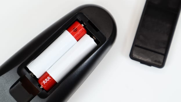 Black TV remote control with AAA alkaline batteries in red and white on a white background. Battery replacement, spare parts. Remote control battery compartment close-up