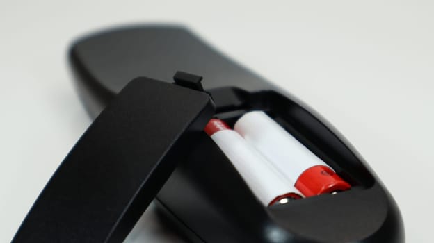 Black TV remote control with AAA alkaline batteries in red and white on a white background. Battery replacement, spare parts. Remote control battery compartment close-up