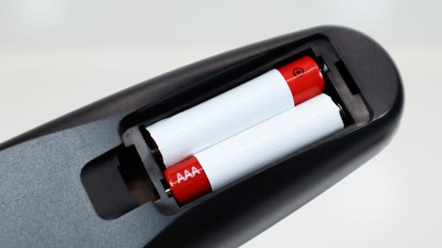 Black TV remote control with AAA alkaline batteries in red and white on a white background. Battery replacement, spare parts. Remote control battery compartment close-up