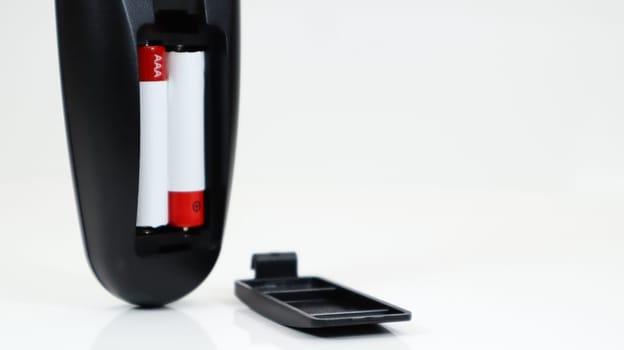 Black TV remote control with AAA alkaline batteries in red and white on a white background. Battery replacement, spare parts. Remote control battery compartment close-up. Copy space.