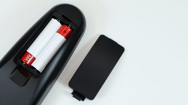 Black TV remote control with AAA alkaline batteries in red and white on a white background. Battery replacement, spare parts. Remote control battery compartment close-up. Copy space