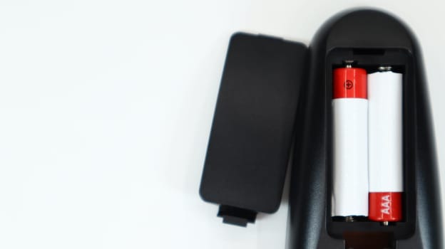 Black TV remote control with AAA alkaline batteries in red and white on a white background. Battery replacement, spare parts. Remote control battery compartment close-up. Copy space