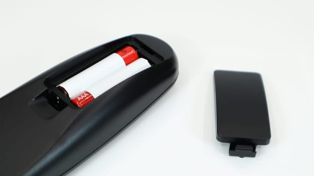 Black TV remote control with AAA alkaline batteries in red and white on a white background. Battery replacement, spare parts. Remote control battery compartment close-up