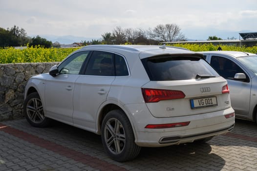 Gaziveren Cyprus -15.02.2025 A stylish white SUV rests near a stone wall, surrounded by vibrant greenery and a bright blue sky filled with clouds.
