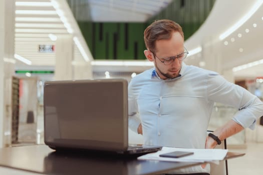 Man holds on to his back, suffers from lower back pain, working on computer. Sedentary work office worker.