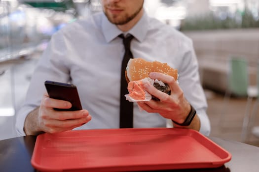 Office worker in blue shirt and black tie snacking on burger in mall, surfing the internet in a mobile while eating. Rush, social media addiction