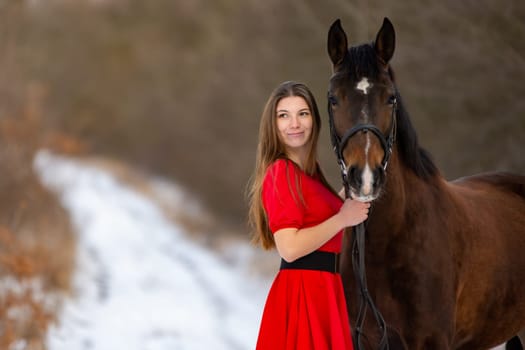 Portrait of a beautiful girl in a red dress standing with a horse on the background of a winter road