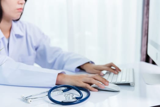 Asian beautiful female doctor smiling wearing doctor's uniform with stethoscope typing on keyboard desktop computer looking to monitor on the desk at hospital office, Medical healthcare concept