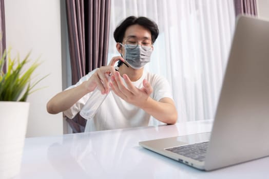 Asian business young handsome man glasses working from home office he quarantines disease coronavirus or COVID-19 wearing protective mask and cleaning hands with sanitizer gel on front laptop computer