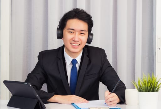 Work from home, Asian young businessman wear suit video conference call or facetime he smiling sitting on desk using smart digital tablet computer and write paper notebook information at home office