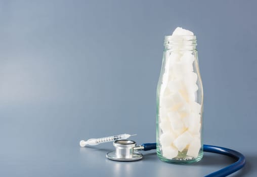 Glass bottle full of white sugar cube sweet food ingredient and doctor stethoscope isolated gray background, health high blood risk of diabetes and calorie intake concept and unhealthy drink