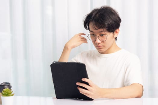 Happy Asian young business handsome man work from home office wear glasses, t-shirt comfortable he smiling and using a black modern smart digital tablet computer to read email or learning on desk