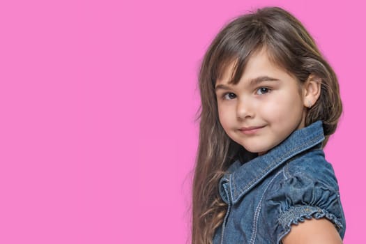Smiling cute tanned long haired little girl wears  denim costume is posing on pink background.