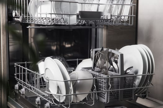 Front view of open automatic stainless steel built-in fully integrated top control dishwasher range machine with clean utensils, cutlery, glasses, dishes, plates inside in modern home kitchen