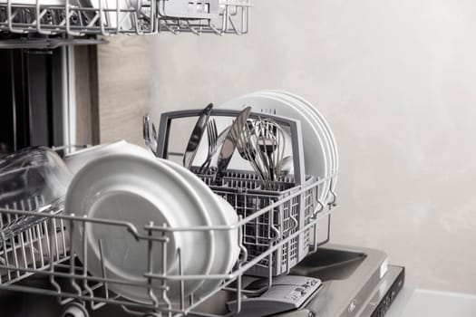 Front view of open automatic stainless steel built-in fully integrated top control dishwasher range machine with clean utensils, cutlery, glasses, dishes, plates inside in modern home kitchen