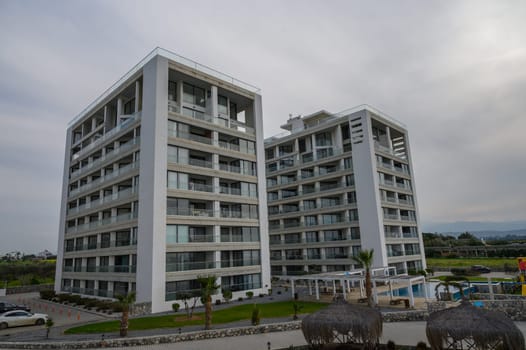 Two modern apartment buildings feature unique designs, set against a cloudy sky with palm trees and greenery.