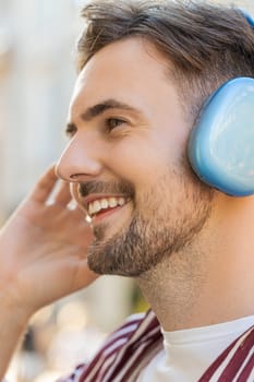 Portrait face of happy relaxed bearded young adult man in wireless headphones choosing listening favorite energetic disco rock n roll music dancing outdoor. Guy standing on urban city street. Vertical