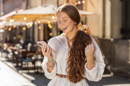 Teenager redhead girl use mobile smartphone celebrating win good message news, lottery jackpot victory, giveaway online outdoors. Happy young woman tourist walking in urban city street. Lifestyles