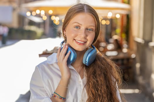 Joyful relaxed overjoyed redhead teenager girl choosing, listening favorite energetic disco rock n roll music dancing outdoors. Child taking off wireless headphones looking at camera on city street