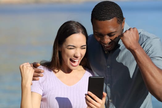 Excited interracial couple checking smart phone on the beach on summer vacation
