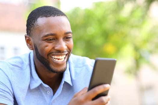 Happy man with black skin checking smart phone laughing sitting in a park
