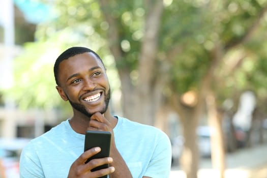 Happy man with black skin thinking holding smart phone in a park