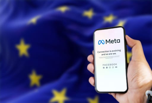 Rome, Italy, Feb 2022: hand holding a phone with the Meta mobile app on the screen and the waving European Union Flag blurred in the background. Meta is the new technology company that owns Facebook