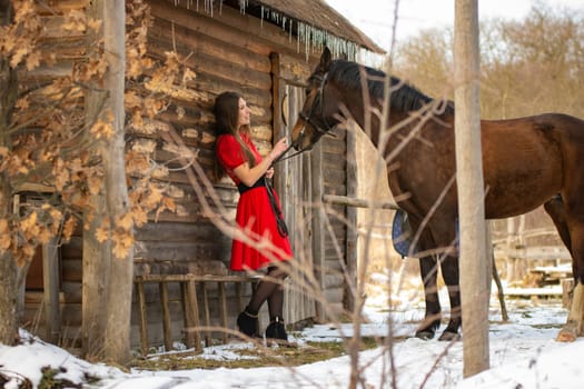A girl in a red dress stands near an old wooden house, a horse stands next to her.