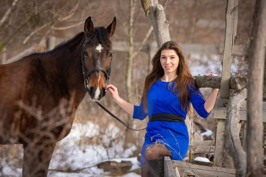 A beautiful girl is sitting on a log against the background of wooden ruins, a horse is standing nearby