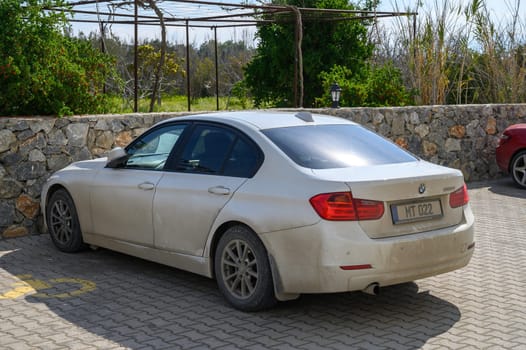 Gaziveren Cyprus -15.02.2025 A dusty BMW rests beside a stone wall, surrounded by greenery on a sunny afternoon.