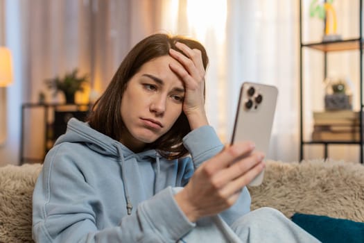 Upset young Caucasian woman sitting on sofa couch disappointed looking at smartphone and placing one hand on forehead expressing deep disappointment due to bad news or losing game. Adult girl at home.
