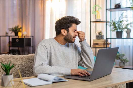 Young Indian man freelancer working on laptop, looking tired, rubbing eyes while sitting at table on sofa in living room at home. Hispanic guy, a workaholic, continues his tasks, clearly exhausted.
