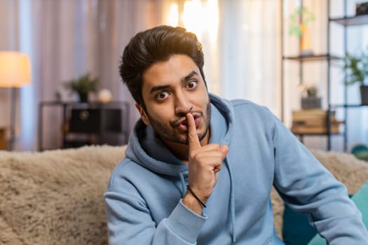 Shh be quiet please. Portrait of young Indian man presses index finger to lips makes silence gesture sign do not tells secret, stop talk gossip, confidential privacy. Guy sitting on sofa at home room.
