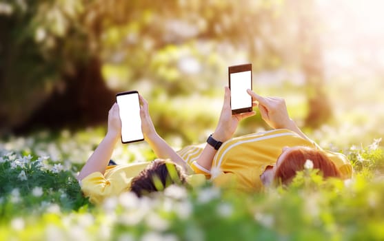 Mother and her son lying down with phones on the meadow in the park. Concept of the family vacations, relationship and using wireless technologies.