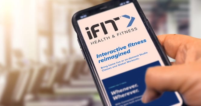 New York, USA, September 2021: hand holding a phone with the iFit mobile app on the screen with a blurred gym in the background during the day. iFIT is a leading health and fitness platform