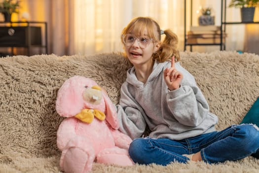 Young teenager girl plays with plush bunny rabbit at home, pretending friendly chat in relaxed mood. Blonde child kid on sofa continues imaginative talk, enjoying gentle bonding with stuffed companion