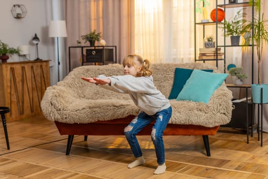 Young teenager girl stands at home performing simple squats and counting one, two, three with growing effort. Child kid sits on sofa finally collapses in laughter, feeling happy after a brief workout