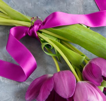 Bouquet of purple tulips with tied engagement or wedding ring and handwritten I LOVE YOU card in envelope on grey table. Valentine's Day celebration, marriage proposal, romantic holiday concept