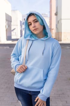 Teenager wearing light blue oversize hoodie and jeans looking into the camera. Blue haired teen girl stays outdoor on city street. Clothing mockup