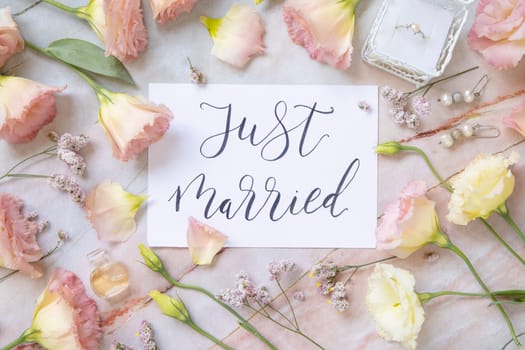 Card with the handwritten text JUST MARRIED surrounded by pink flowers, earrings, petals and parfume flacon top view on a marble table. Romantic concept