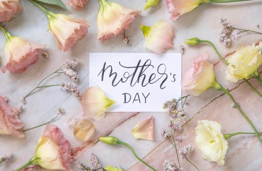 Card with the handwritten text MOTHER'S DAY surrounded by pink flowers, earrings, petals and parfume flacon top view on a marble table. Romantic concept