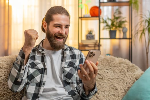 Happy smiling young man sitting at home, scrolling phone receiving good news notification reveals lottery success or game win, sparking joy. Caucasian guy on sofa celebrating success with a big grin