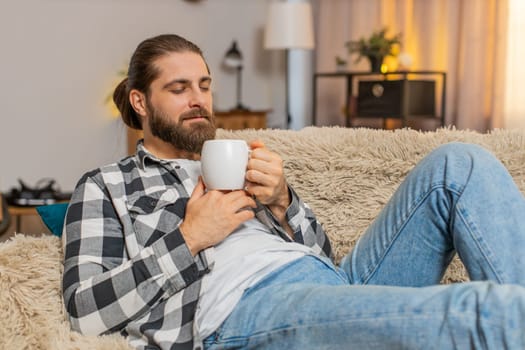 Young man resting lying at home, holding warm cup of coffee or tea with gentle grin and eased shoulders. Caucasian guy on sofa feels soothing quiet, appreciating short pause from busy tasks and noise.